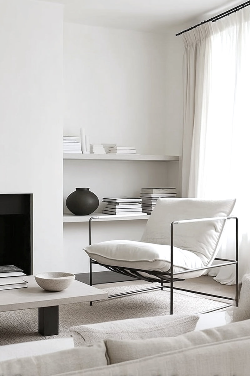 Minimalist white leather lounge chair with slim black iron frame in a bright neutral living room with shelves, linen curtains and a low stone table.