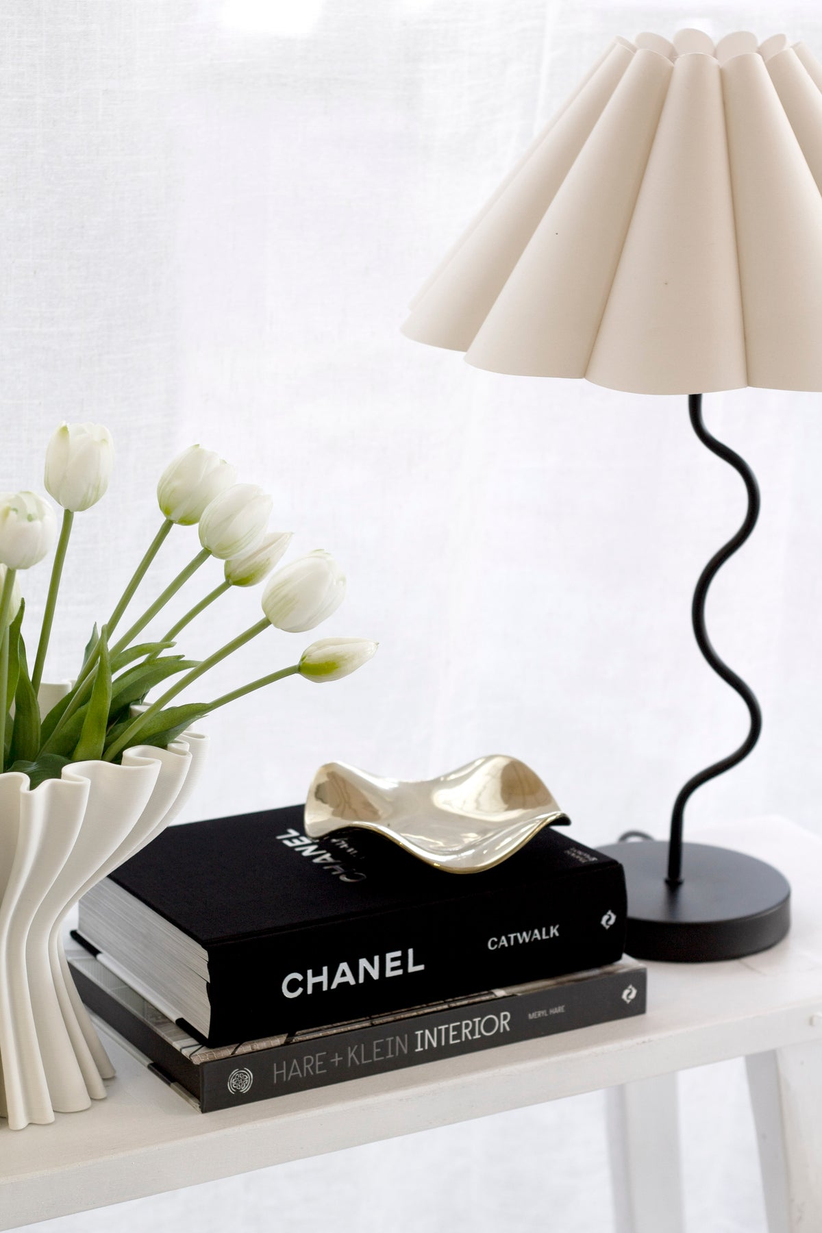 Pale gold wave tray on a stack of black books beside a fluted lamp and white tulips in a ribbed vase