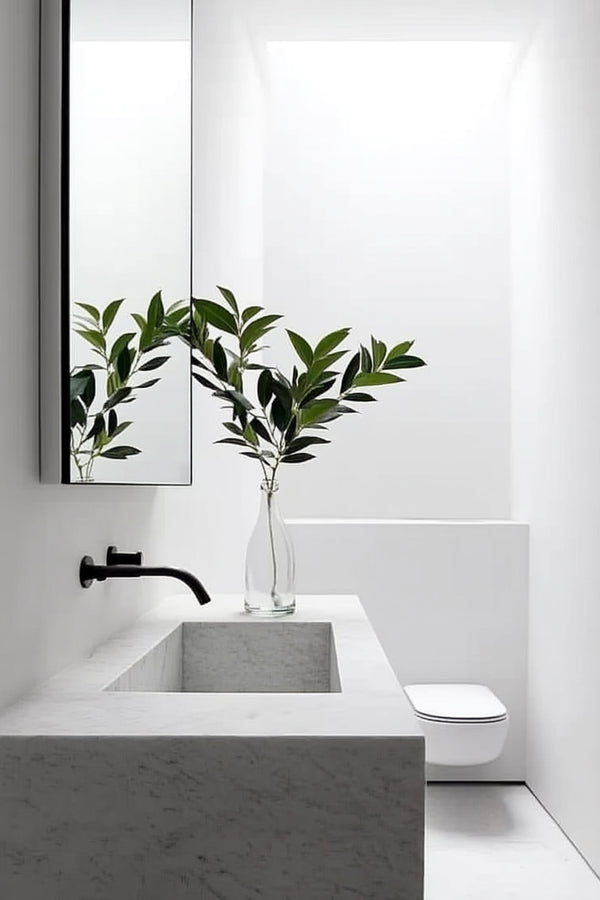 Tall faux green botanical stem styled in a clear vase on a white marble sink in a modern minimalist bathroom with soft natural light and black fittings.