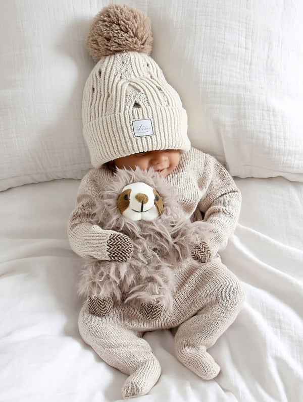 Baby dressed in soft neutral knitwear lying on a white bed, wearing a cream pom-pom beanie and cuddling a fuzzy sloth rattle with herringbone-knit paws and a sweet face.

