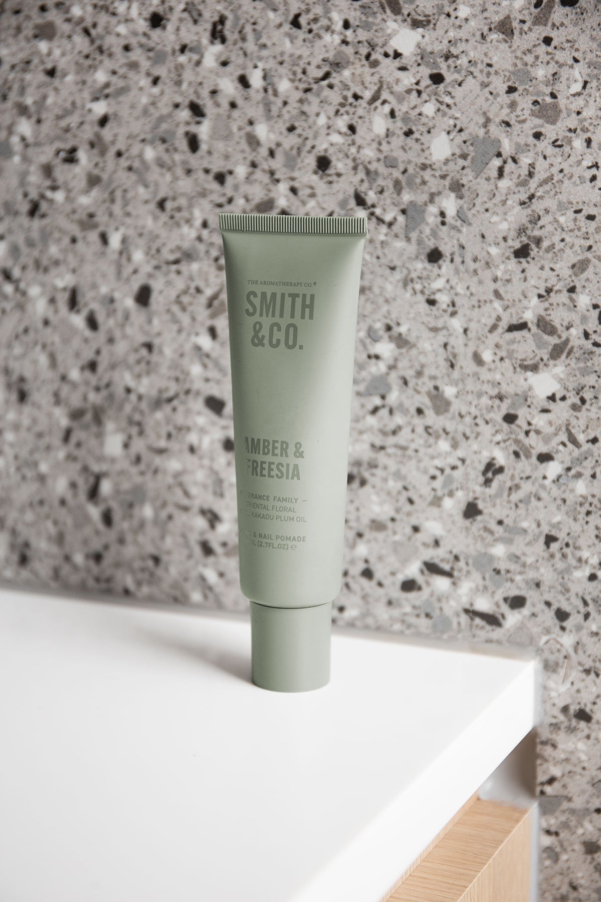 Smith & Co. hand and nail pomade in Amber & Freesia, sage green tube styled on a white vanity shelf against a terrazzo wall