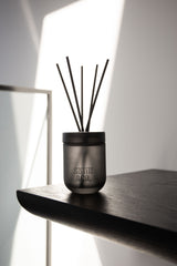 Smith and Co Tabac and Cedarwood reed diffuser in a dark gunmetal frosted glass vessel with black reeds on a dark timber shelf in dramatic natural light against a white wall