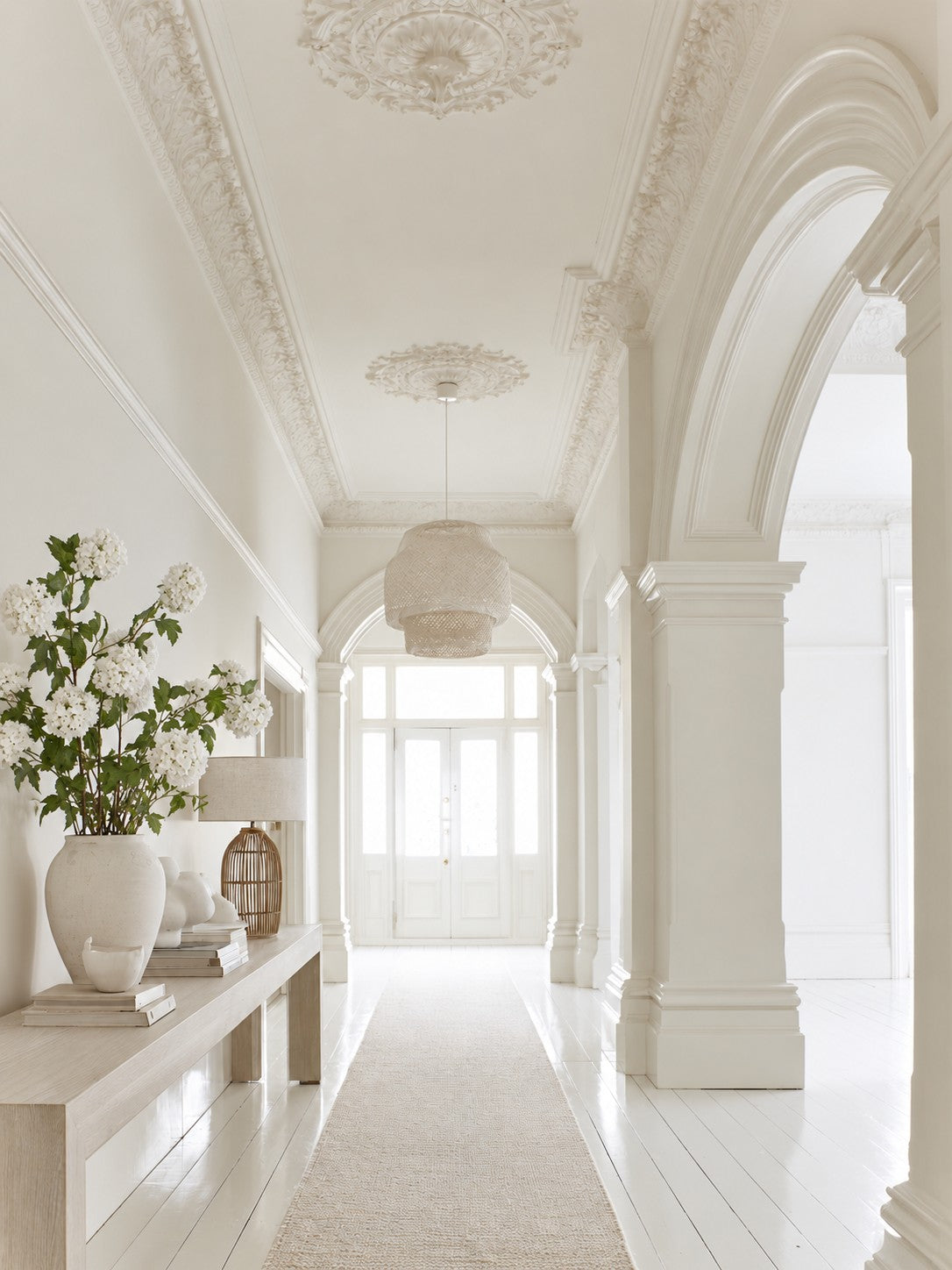 White faux snowball viburnum stems in a stone vase on a hallway console beside a rattan lamp