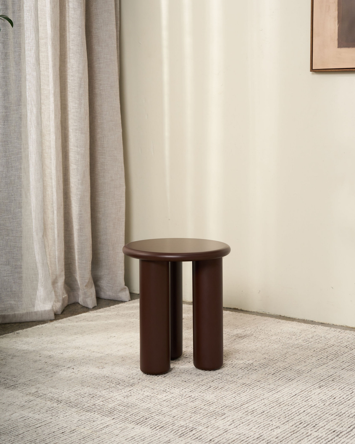 Rouge side table on a pale rug in the corner of a room with linen curtains