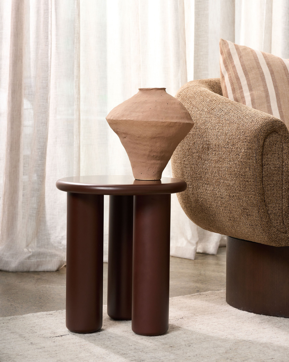 Rouge side table holding a terracotta clay vessel beside a textured armchair