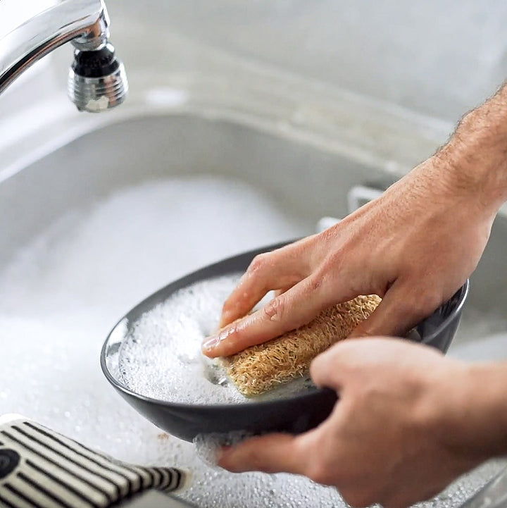 A person using an ECO SCRUBS 2-PACK, a natural option, to wash a dish with a scourer sponge in a sink.