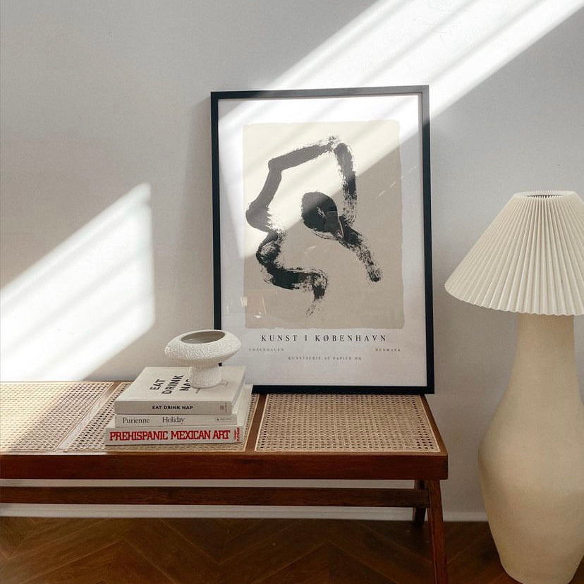 Copenhagen print in a black frame on a rattan bench beside books and a pleated lamp