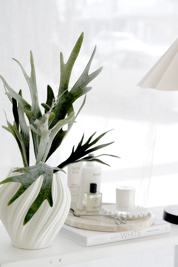 Artificial staghorn fern stem styled in white vase on console with books and lamp; realistic faux greenery for home decor.