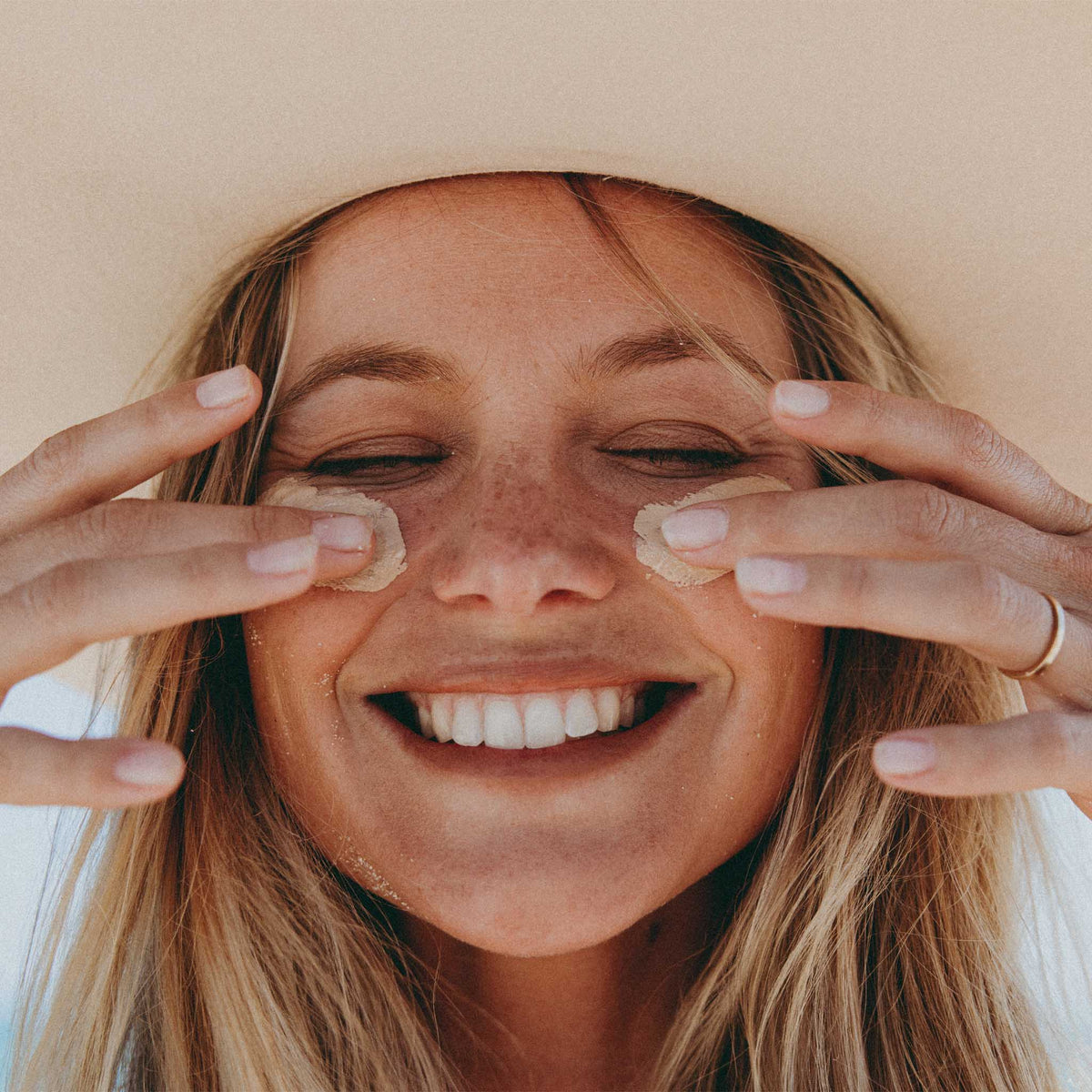 Woman applying natural sticky zinc sunscreen to face at the beach wearing a sun hat