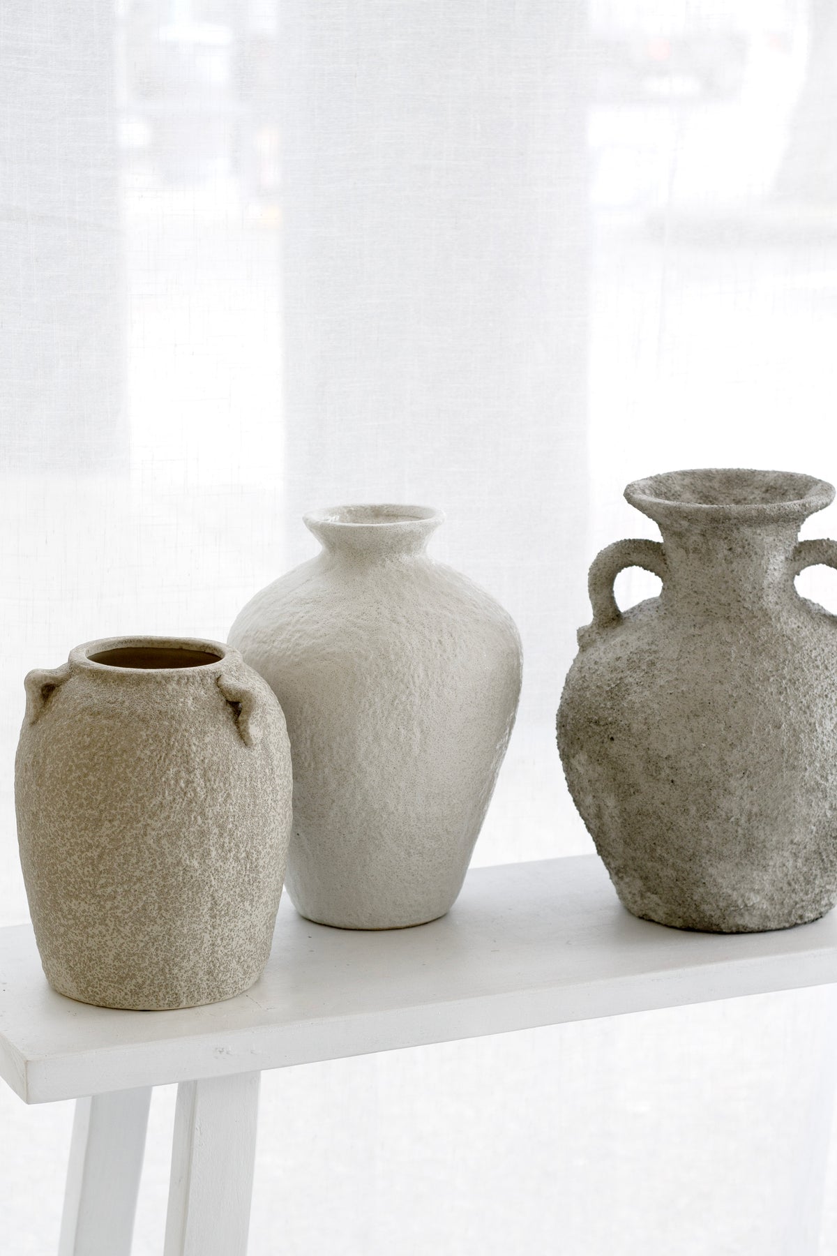 Trio of textured stone vases styled in front of a sheer curtained window, tonal beige palette.
