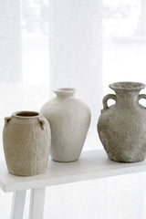 Trio of textured stone vases styled in front of a sheer curtained window, tonal beige palette.