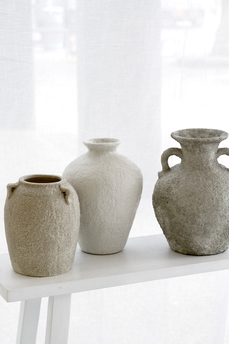 Trio of textured stone vases styled in front of a sheer curtained window, tonal beige palette.