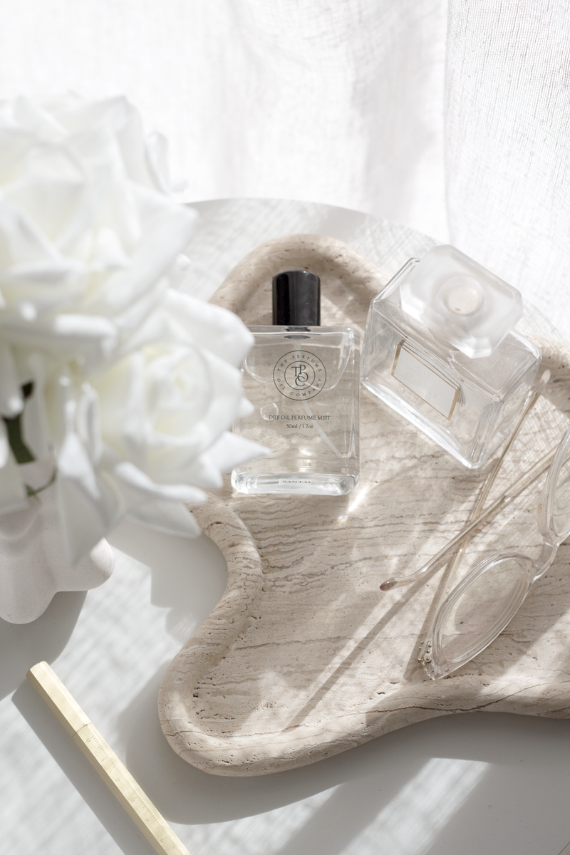 Travertine curved tray with clear perfume bottles and glasses in soft sunlight; neutral “styling with trays” bedside vignette.