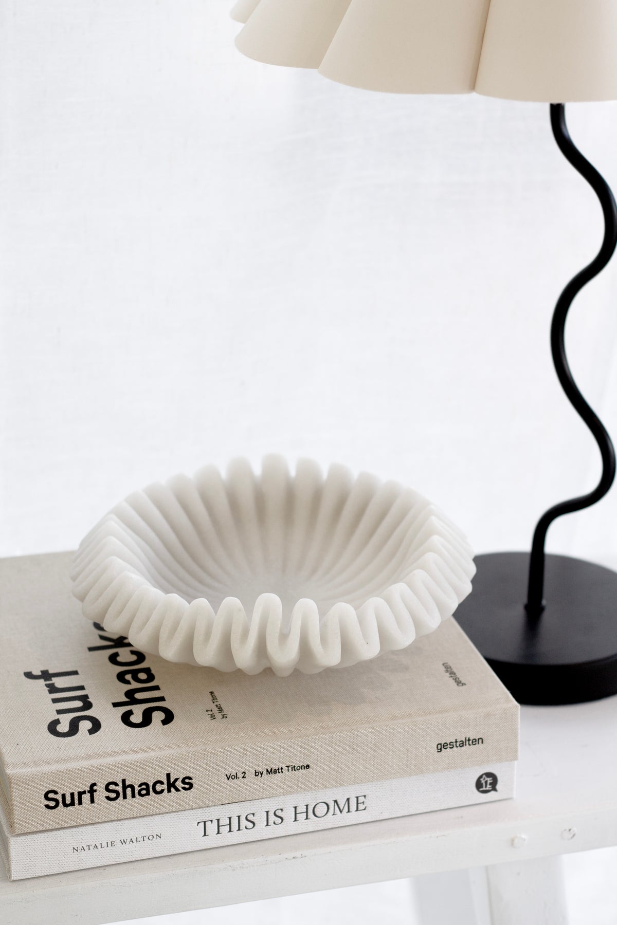 Surf Shacks Volume 2 coffee table book styled with a sculptural white dish and black wavy lamp on a neutral shelf.