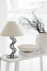 Surge Lamp with ripple-shaped black steel base and linen shade styled on a white console with sculptural decor and modern books.