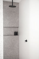 Minimalist shower with terrazzo tiles and matte black rain shower head, featuring two black Tabac & Cedar Wood wash bottles on a tiled ledge.