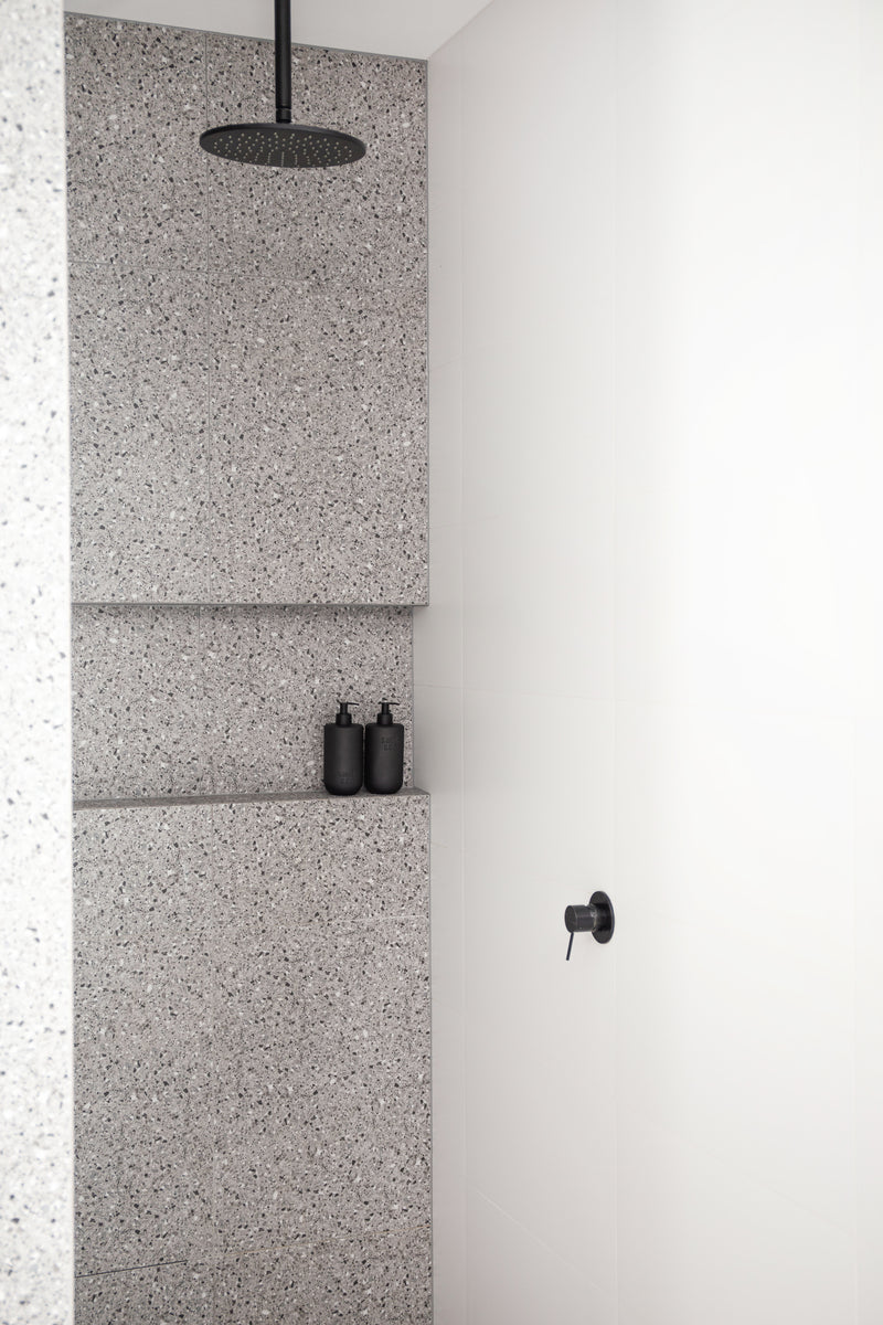 Minimalist shower with terrazzo tiles and matte black rain shower head, featuring two black Tabac & Cedar Wood wash bottles on a tiled ledge.