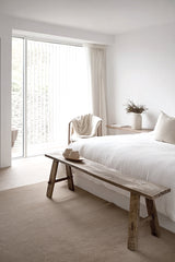 A rustic recycled teak bench styled at the foot of a bed in a minimalist bedroom with soft white bedding, neutral tones, and natural light filtering through sheer curtains.