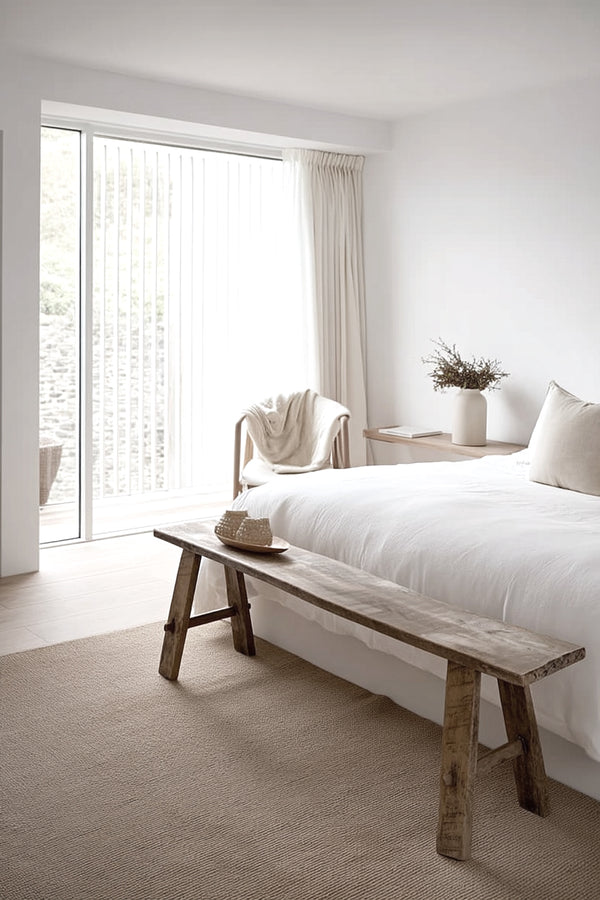 A rustic recycled teak bench styled at the foot of a bed in a minimalist bedroom with soft white bedding, neutral tones, and natural light filtering through sheer curtains.