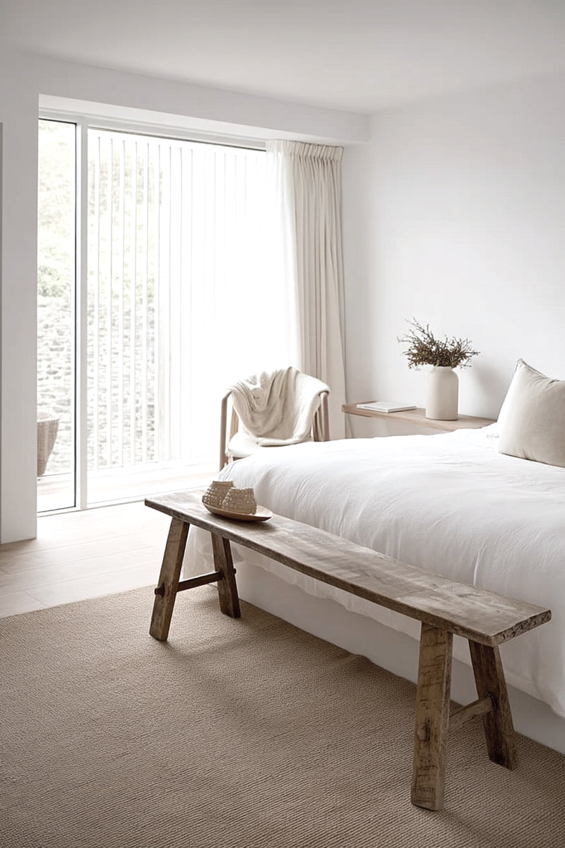 A rustic recycled teak bench styled at the foot of a bed in a minimalist bedroom with soft white bedding, neutral tones, and natural light filtering through sheer curtains.