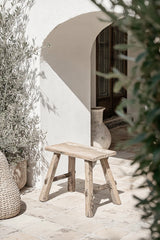 Small rustic bench handcrafted from recycled teak wood, styled outdoors against a white stucco wall with textured stone tiles, surrounded by woven baskets and greenery, evoking a coastal Mediterranean aesthetic.