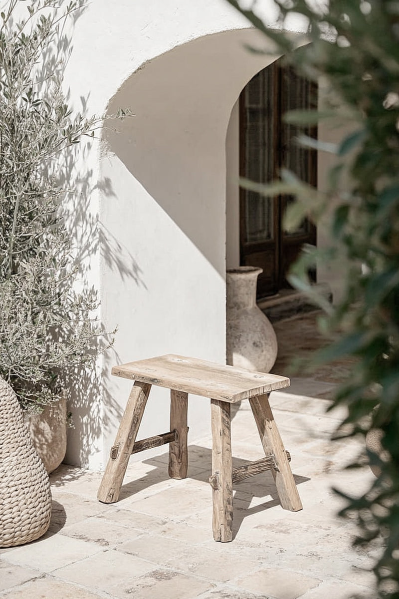Small rustic bench handcrafted from recycled teak wood, styled outdoors against a white stucco wall with textured stone tiles, surrounded by woven baskets and greenery, evoking a coastal Mediterranean aesthetic.