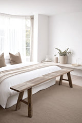 A long, weathered teak bench sits at the foot of a bed in a minimalist bedroom with soft neutral tones, sheer curtains, and natural textures.