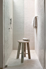 A rustic round teak stool in a minimalist tiled shower, styled with folded white towels and soft natural light.