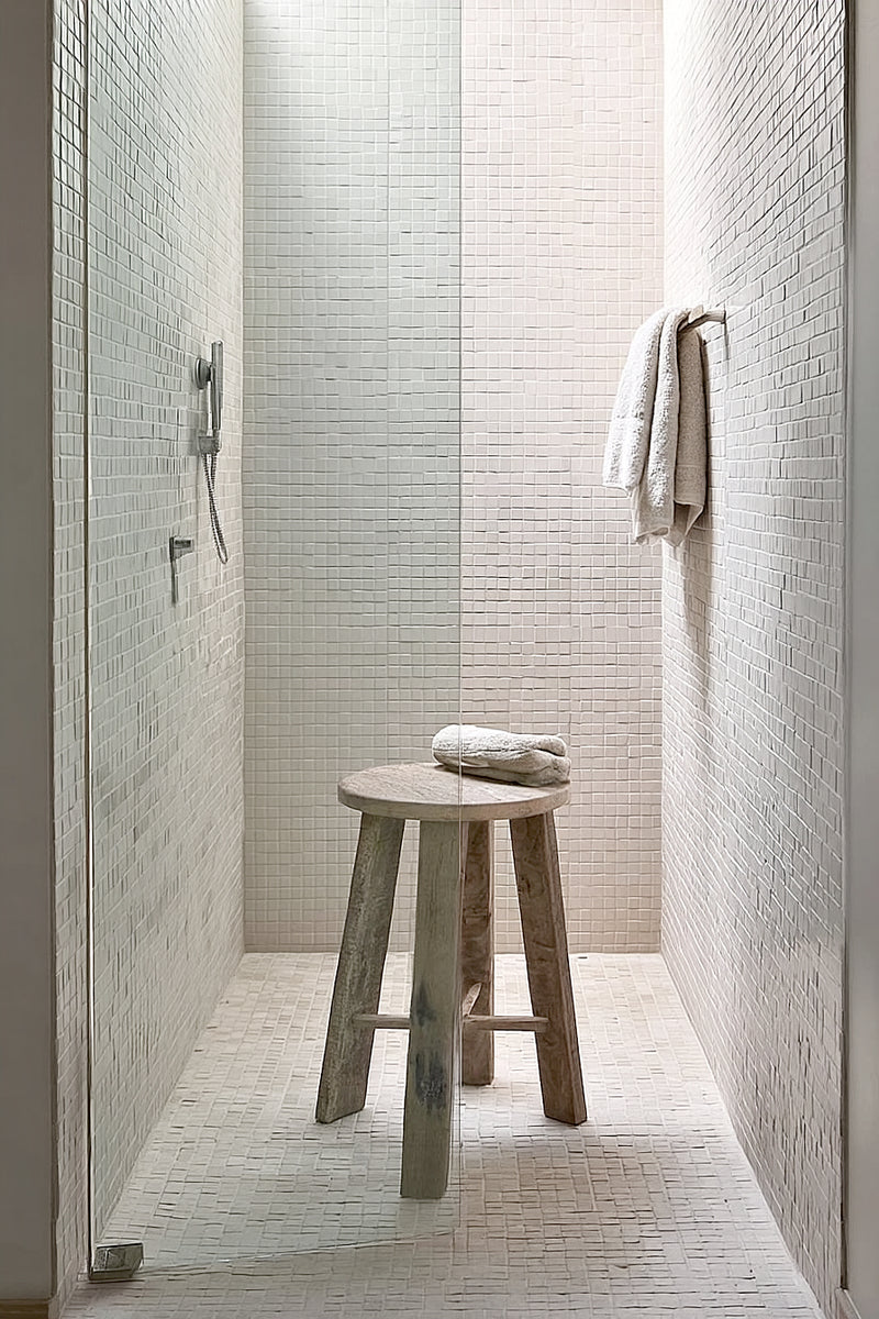A rustic round teak stool in a minimalist tiled shower, styled with folded white towels and soft natural light.