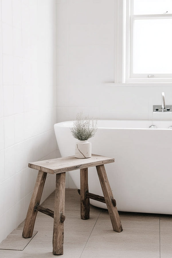 A rustic recycled teak bench styled in a minimalist white bathroom, beside a freestanding tub with a small marble planter on top.