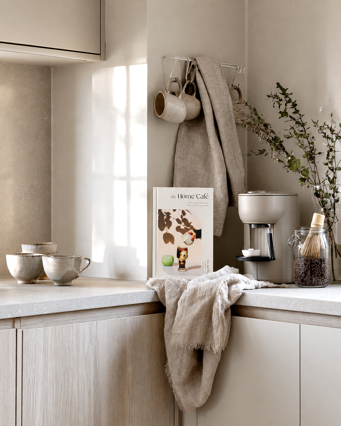 The Home Café recipe book by Asia Lui Chapa propped on a neutral kitchen bench beside speckled ceramic mugs, a matcha whisk in a jar of coffee beans, a coffee machine and a vase of eucalyptus, styled in a soft coastal Scandi palette.