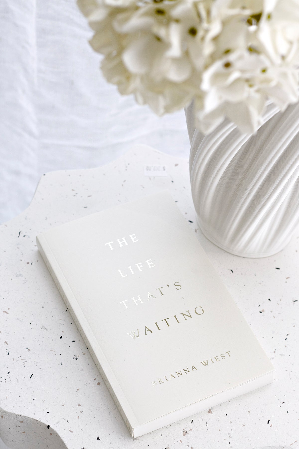 The Life That's Waiting on a terrazzo side table beside a white ribbed vase of white flowers