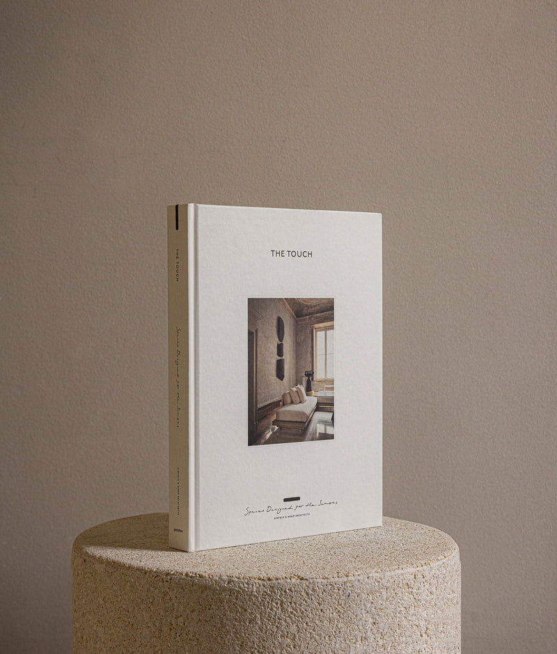 Hardcover design book The Touch: Spaces Designed for the Senses displayed on a textured stone pedestal, minimalist neutral background.