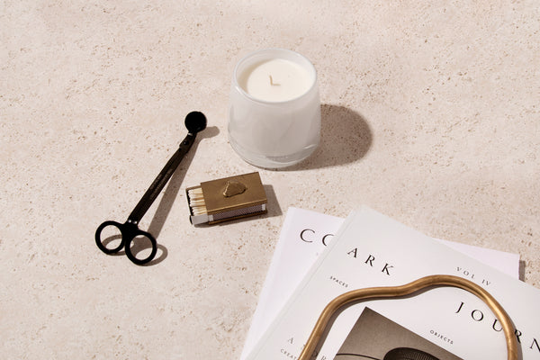 White candle on travertine with black wick trimmer, brass matchbox and design magazines in warm light.