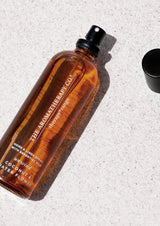 Amber Therapy home and linen spray bottle labelled Coconut & Water Flower lying on a pale stone surface with its black spray lid off to the side in bright sunlight.
