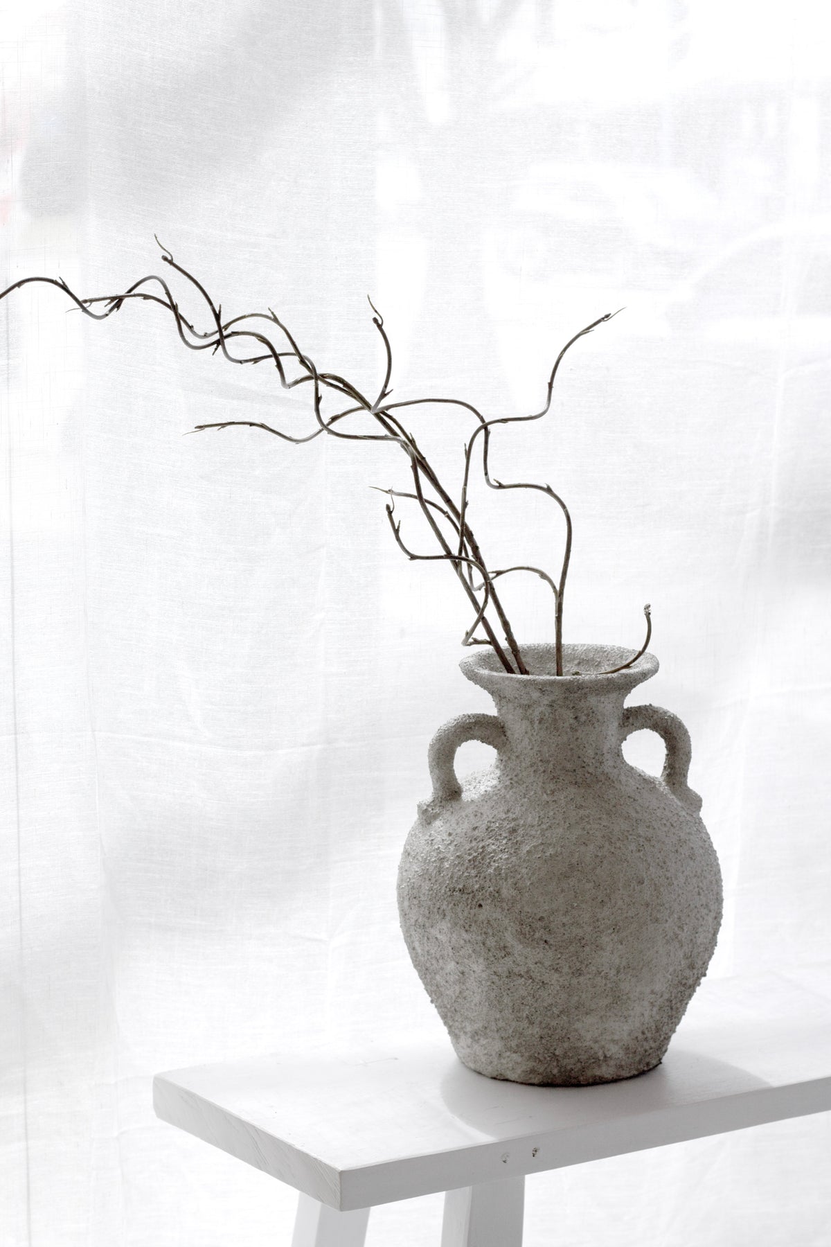 Thira Vase styled with a branch on a white plinth in soft daylight