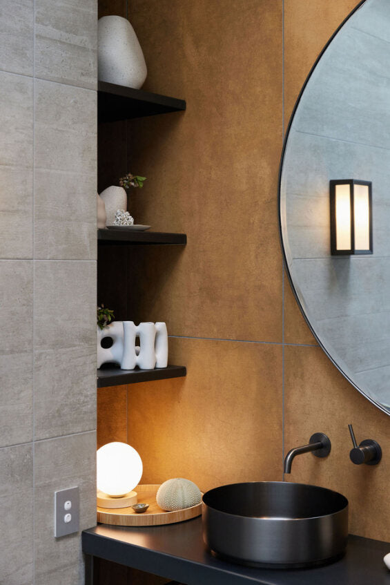 Yuri Table Lamp glowing on a dark bathroom vanity beside a round mirror and a black basin