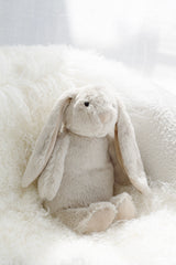 Neutral plush bunny soft toy in biscuit colour styled on white textured cushion
