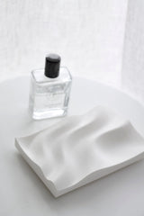 White wavy 3D-printed jewellery tray styled on a minimal white surface beside a clear glass perfume bottle in soft natural light.