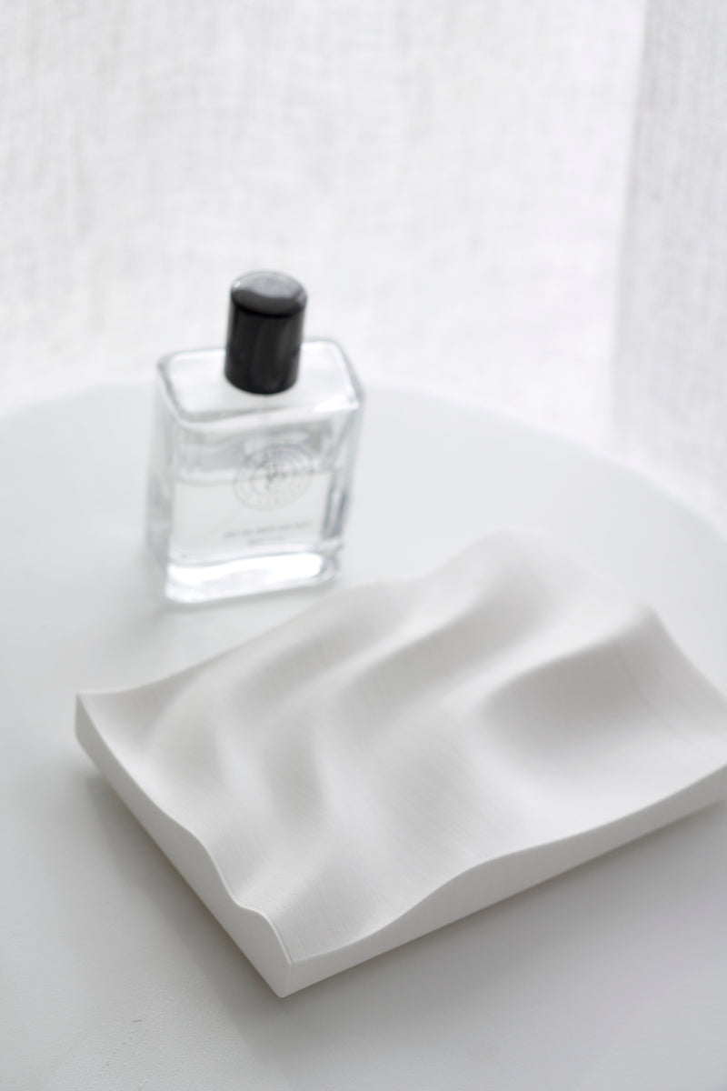 White wavy 3D-printed jewellery tray styled on a minimal white surface beside a clear glass perfume bottle in soft natural light.