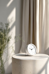 Minimalist Karlsson Tinge Alarm Clock in white with a foldable stand, styled on a round stone pedestal in soft natural light with neutral curtains and greenery in the background.