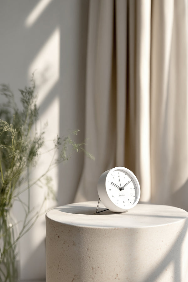 Minimalist Karlsson Tinge Alarm Clock in white with a foldable stand, styled on a round stone pedestal in soft natural light with neutral curtains and greenery in the background.