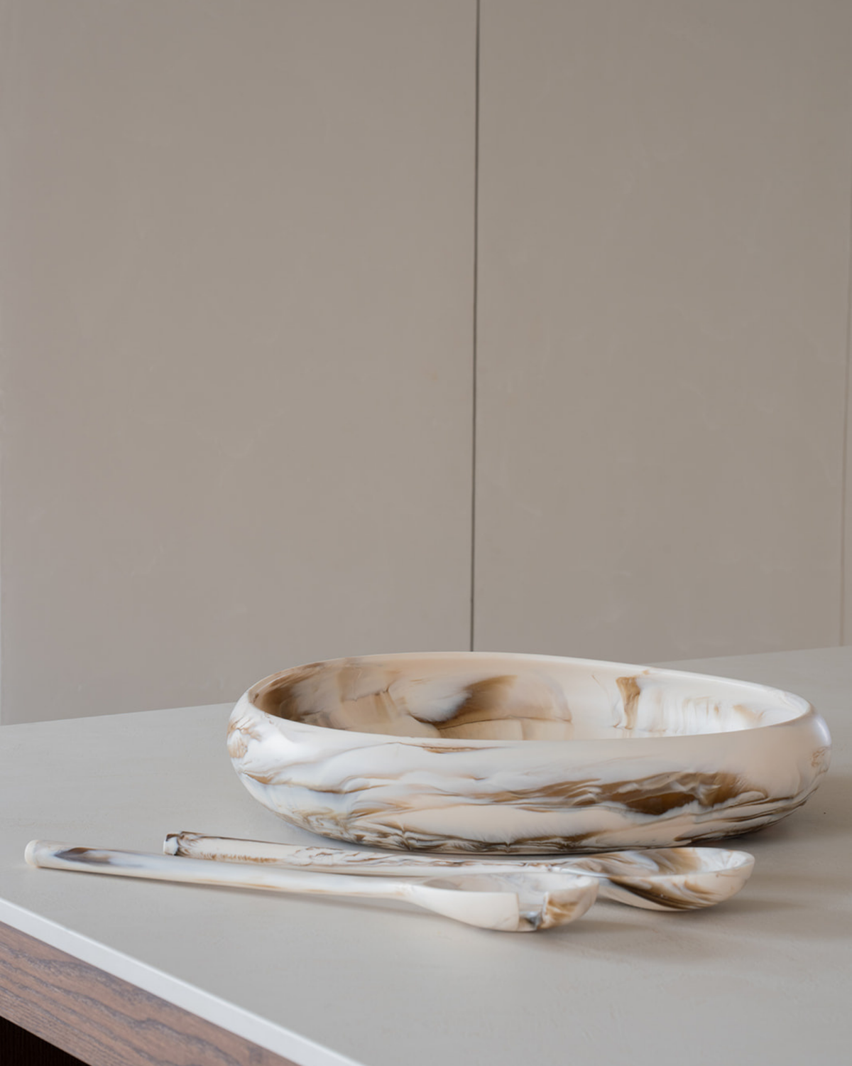 Tiramisu salad servers lying beside a large cream marbled resin bowl on a white benchtop