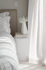 White ribbed polyurethane stool styled as bedside table beside neutral bed and sheer curtains