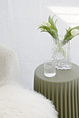 Ribbed olive-green Tom polyurethane stool used as a side table, styled with a clear textured glass vase of white tulips and a ribbed glass tumbler beside a boucle chair in a bright neutral room