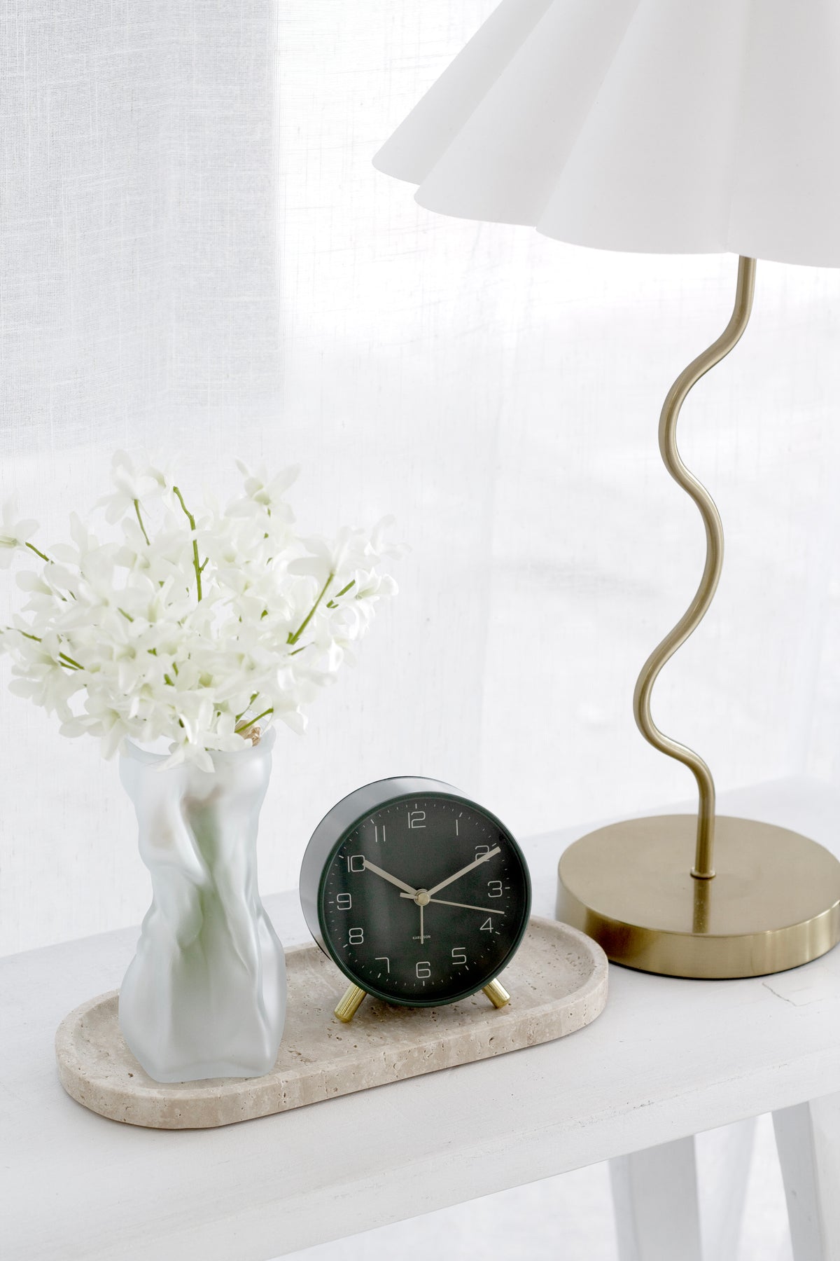Travertine line tray with white vase of flowers and small black clock beside a brass lamp on a white console.