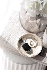 Natural travertine round tray with warm beige veining styled with jewellery and a smart watch on a book stack beside a glass vase of white roses.
