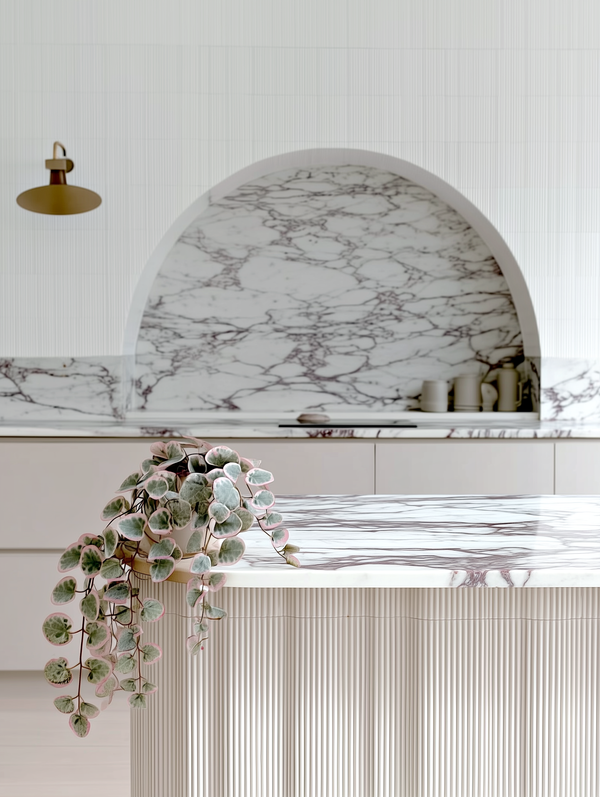 Artificial variegated string of hearts plant with heart-shaped green and pink leaves trailing across a marble kitchen island in a modern light-filled space.