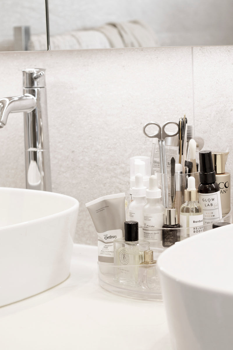 Clear rotating Umbra Cascada cosmetic organizer on a bathroom vanity, filled with skincare bottles, droppers, sprays and makeup brushes between two white basins.