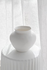 Venus Vase in matte white ceramic styled on a pedestal with soft natural light filtering through sheer curtains.